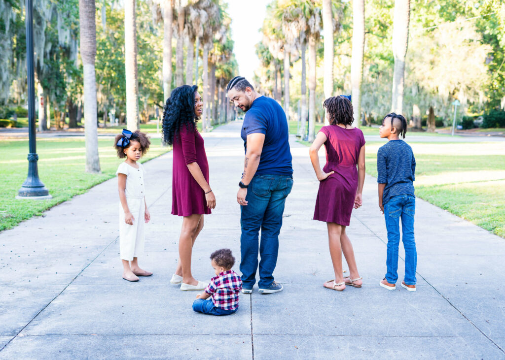 family of 6 walking down palm lined sidewalk looking back at toddler sitting on ground