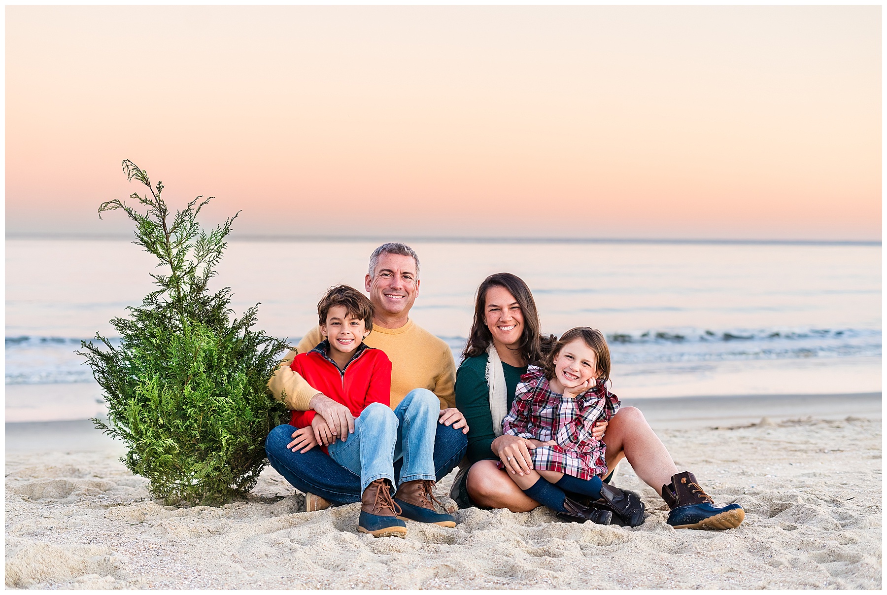 family of four sitting on the beach with a Christmas tree having a family photo taken