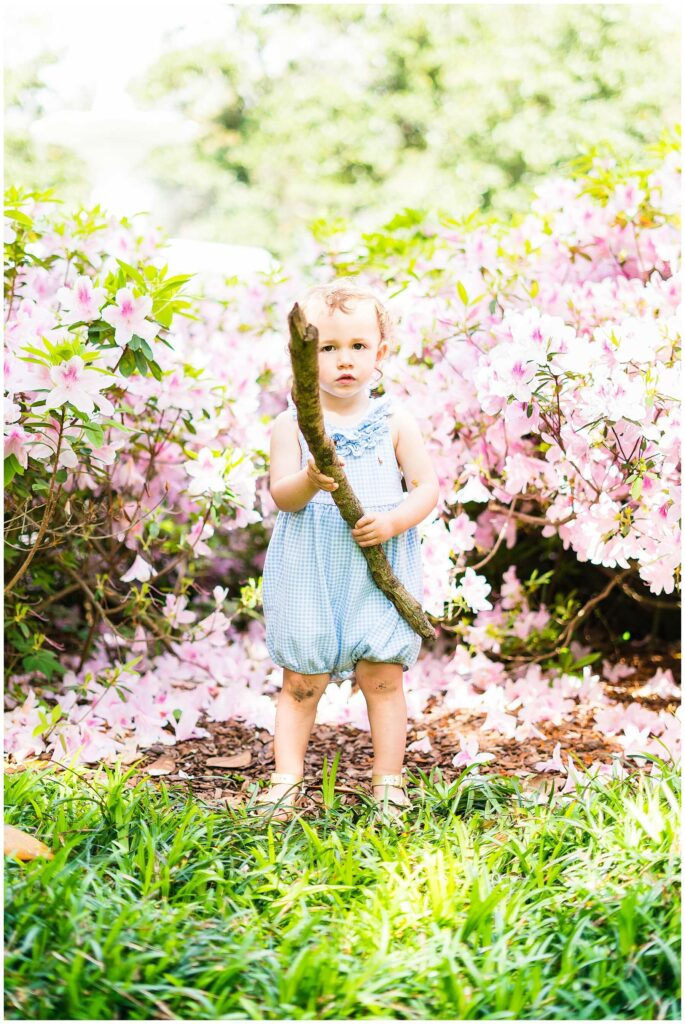 little girl dressed up with dirty knees holding a large stick standing in the azaleas at Forsyth Park