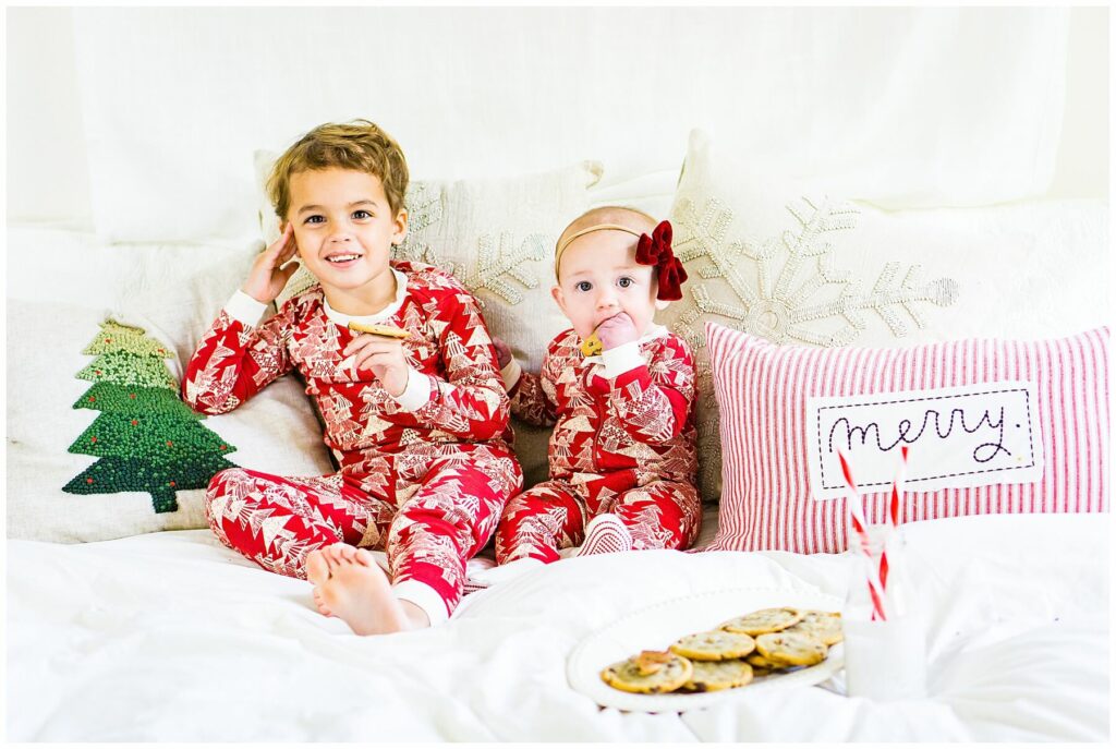 milk and cookie mini sessions two kids sitting on a bed in Christmas pajamas eating cookies