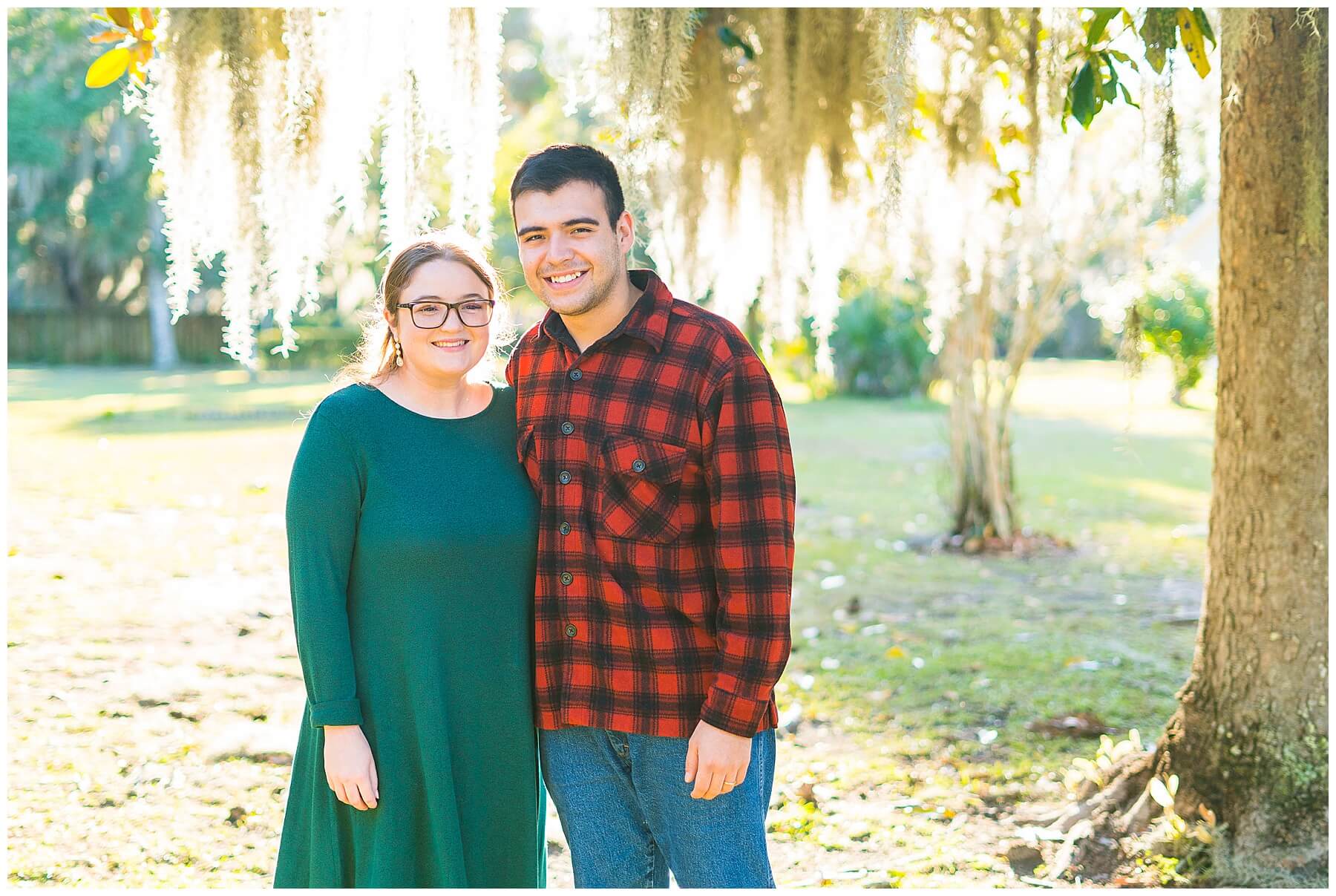 young couple dressing in Christmas green and red buffalo plaid smiling at camera with light flowing through Spanish moss