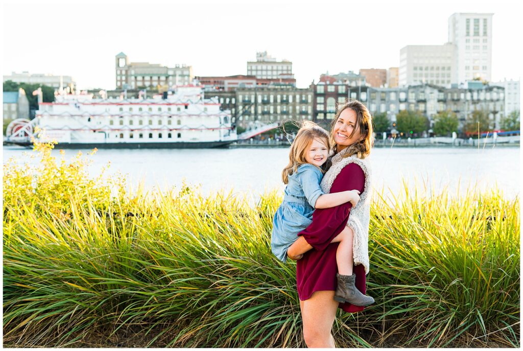 woman in maroon dress holding a girl in blue dress with downtown savannah In background
