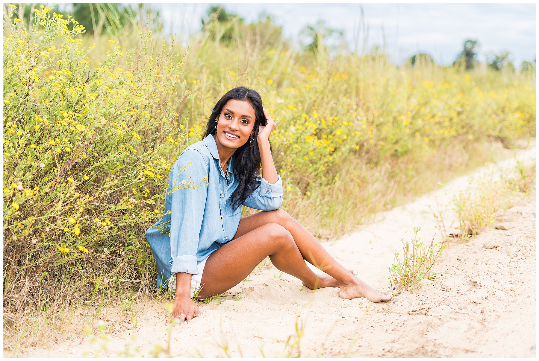 high school senior girl sitting on the side of a dirt road with yellow flowers behind her
