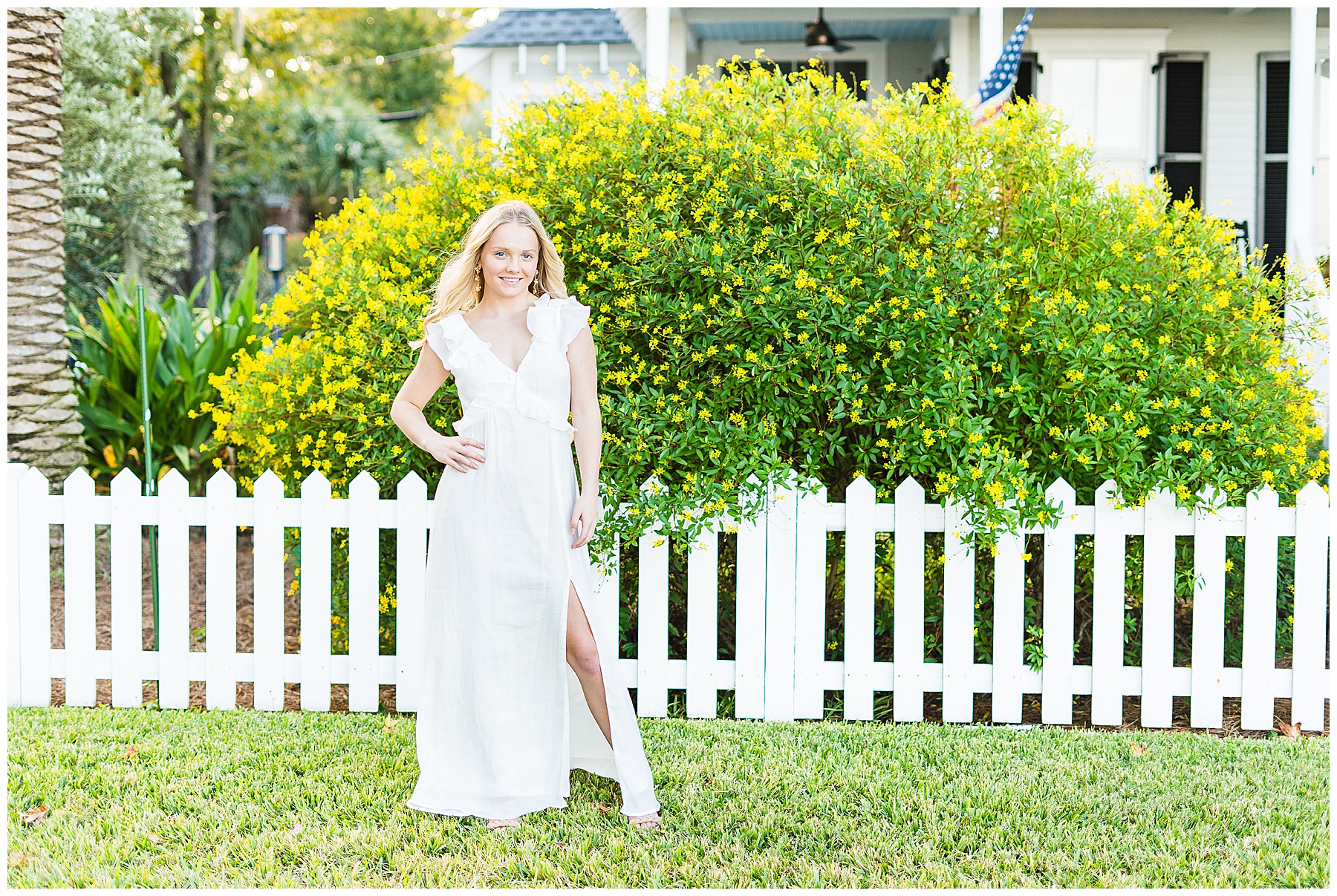 savannah senior standing in a white dress in front of a white picket fence and bush with yellow flowers