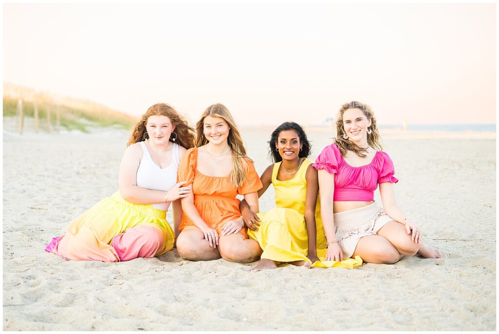 group of high school senior girls wearing various outfits in pinks, oranges, and yellows sitting in the sand on Tybee Island