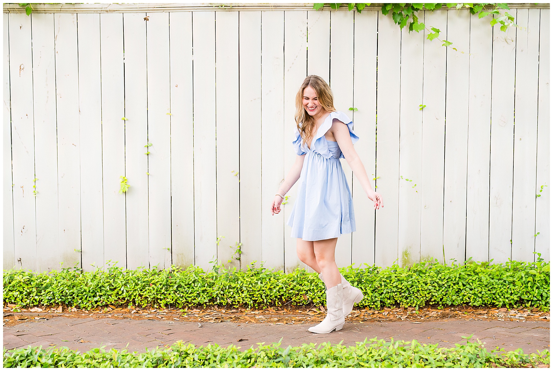 high school senior girl wearing a pale blue dress and cowboy boots walking in front of a white fence in downtown savannah Georgia