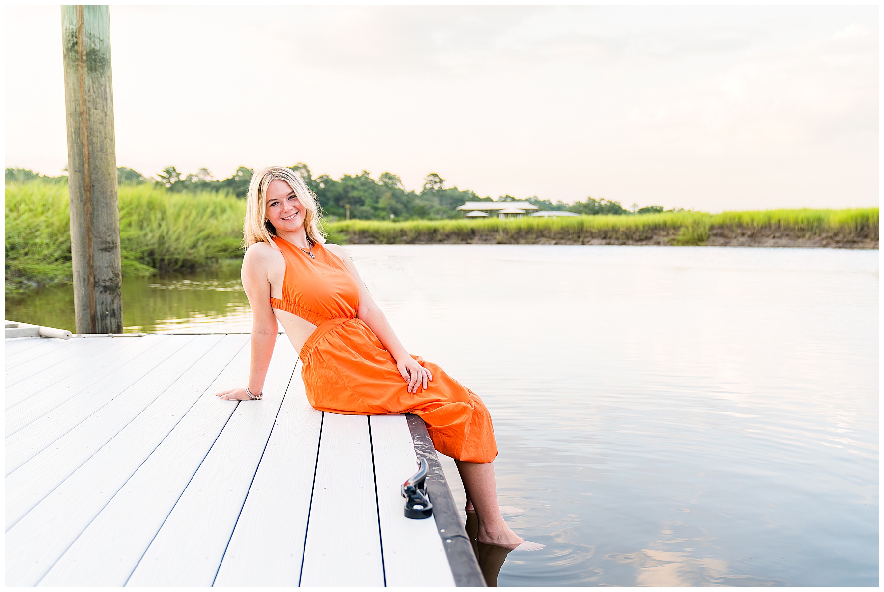 photo of a blonde girl wearing an orange maxi dress sitting on a dock with her feet in the water