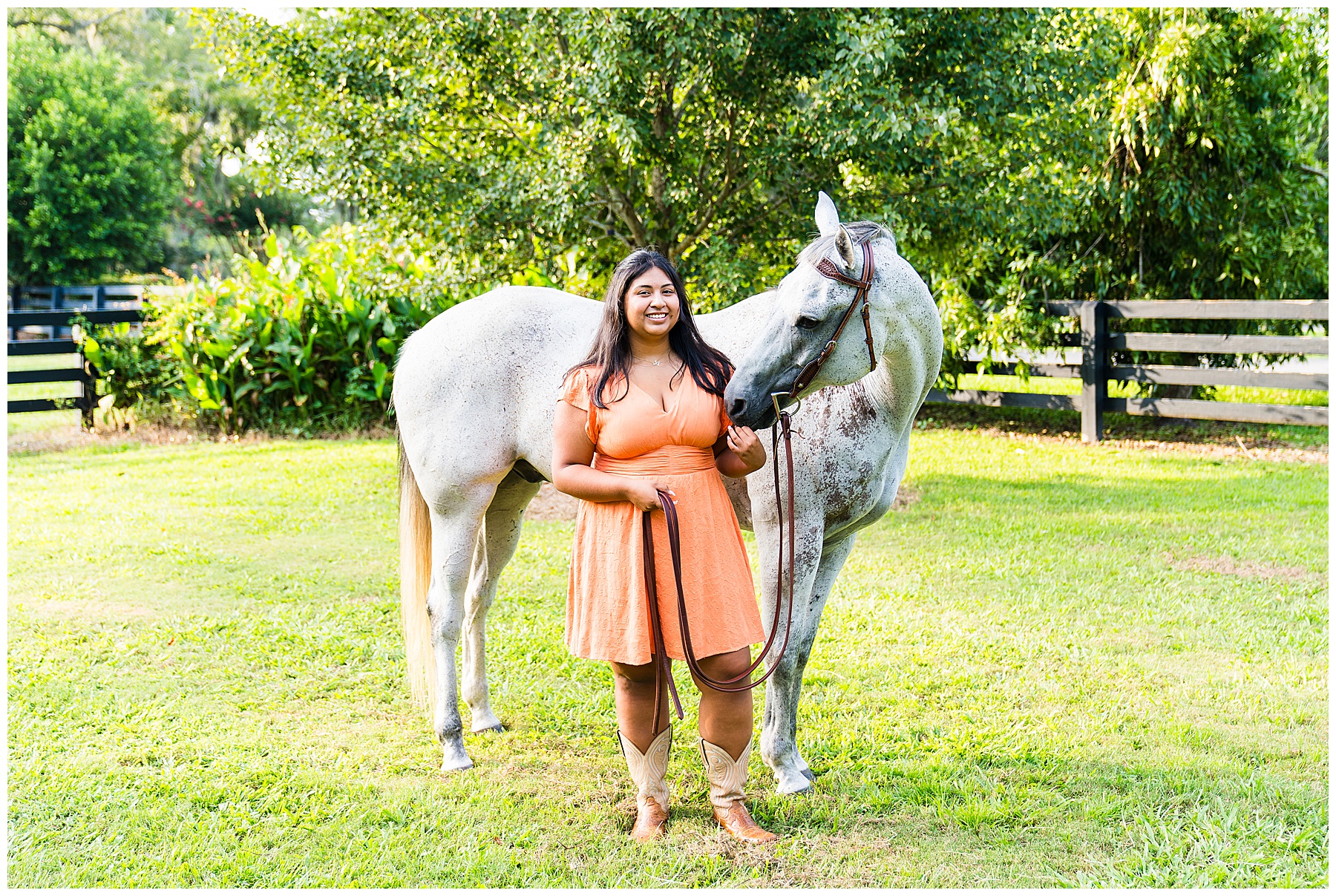 girl wearing an orange dress standing in a grassy field with a grey horse wearing a bridle