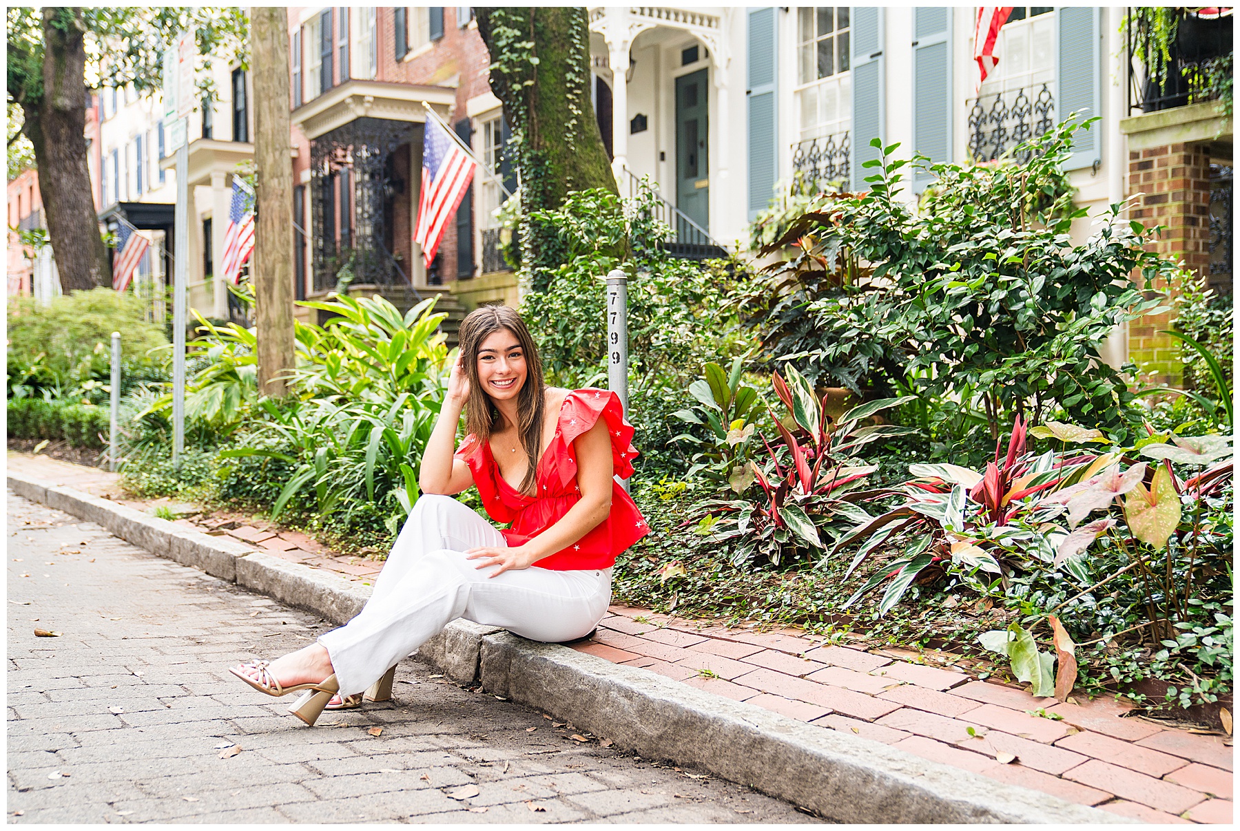 a brunette girl wearing a pair of white pants and a red top sitting on the curb having her senior pictures taken on downtown Savannah's Jones Street