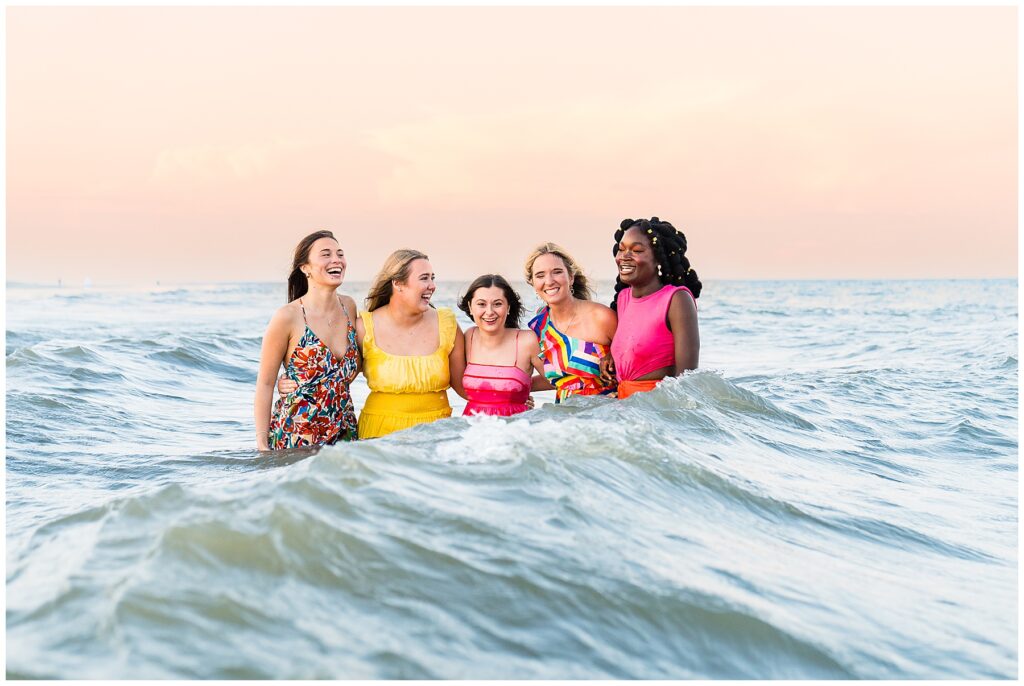 group of high school senior girls wearing bold and bright dresses standing in the ocean at sunset