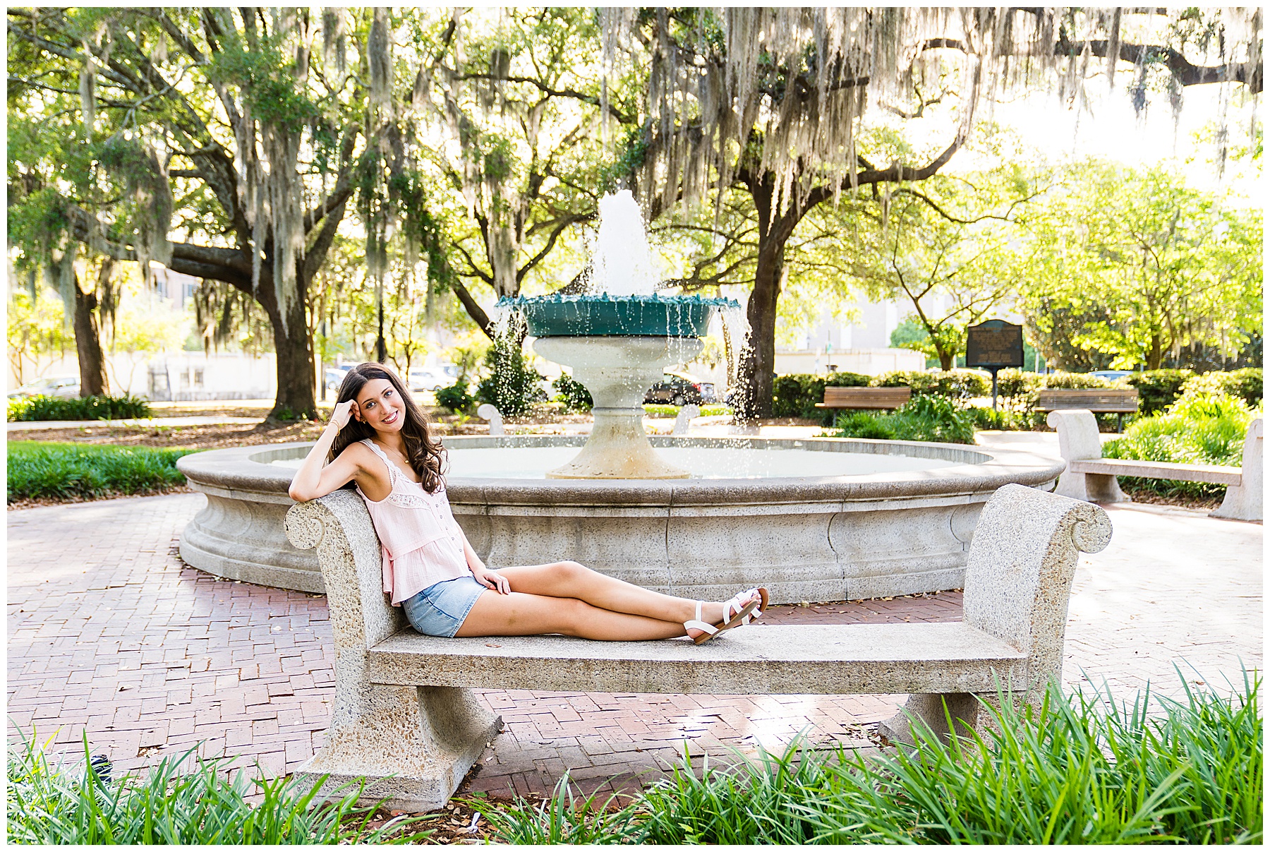 This image captures a dreamy and relaxed senior portrait set in one of Savannah’s iconic squares, filled with Southern charm and timeless beauty. The young woman is lounging on a stone bench in front of a classic fountain with water softly cascading into the basin behind her. She’s wearing a light pink, lace-trimmed sleeveless top, denim shorts, and white strappy sandals—an outfit that feels fresh, summery, and effortlessly stylish. Her long hair falls in soft waves over her shoulder as she leans her head against her hand and smiles sweetly at the camera. Surrounding her are vibrant green plants, red brick pathways, and grand oak trees draped in Spanish moss, creating a romantic and serene atmosphere. The lighting is warm and golden, possibly late afternoon, casting a soft glow across the scene and enhancing the magical feel of the historic park setting.