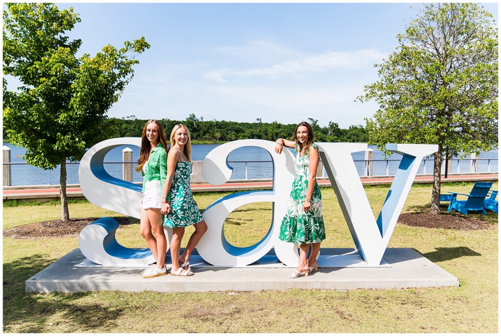 This image features three young women posing in front of large sculptural letters spelling out “SAV,” likely short for Savannah. They are outdoors on a sunny day, standing on a concrete base surrounded by grass, trees, and a riverfront promenade in the background. Two of the women are standing in front of the "S" and "a," while the third leans casually on the "V." They are all dressed in summery outfits featuring green and white tones, including floral patterns and light fabrics, which coordinate beautifully with the natural surroundings. The setting feels cheerful and vibrant—perfect for senior photos or a fun girls’ day out. The relaxed, happy vibe and the bright lighting give the image a fresh, youthful energy.