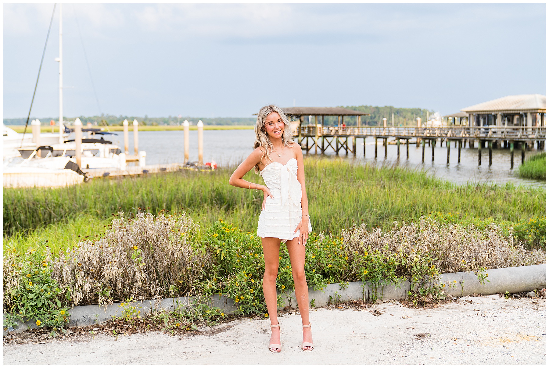 This image captures a senior portrait with a beautiful waterfront backdrop, blending elegance with a coastal, natural setting: The senior stands confidently in the center of the frame, smiling softly with one hand on her hip and the other resting casually by her side. She’s wearing a strapless white textured romper with a large front bow and tiered detailing. Her light pink heeled sandals add a soft pop of color and sophistication to the look. Behind her is a lush mix of green marsh grasses and flowering plants, transitioning to a calm body of water with anchored boats and wooden piers. On the right, a long dock leads to rustic buildings on stilts, enhancing the coastal charm of the scene. The lighting is soft and flattering, likely captured in golden hour, giving her skin and hair a warm, natural glow. The cloudy sky adds depth without overpowering the bright, cheerful mood.