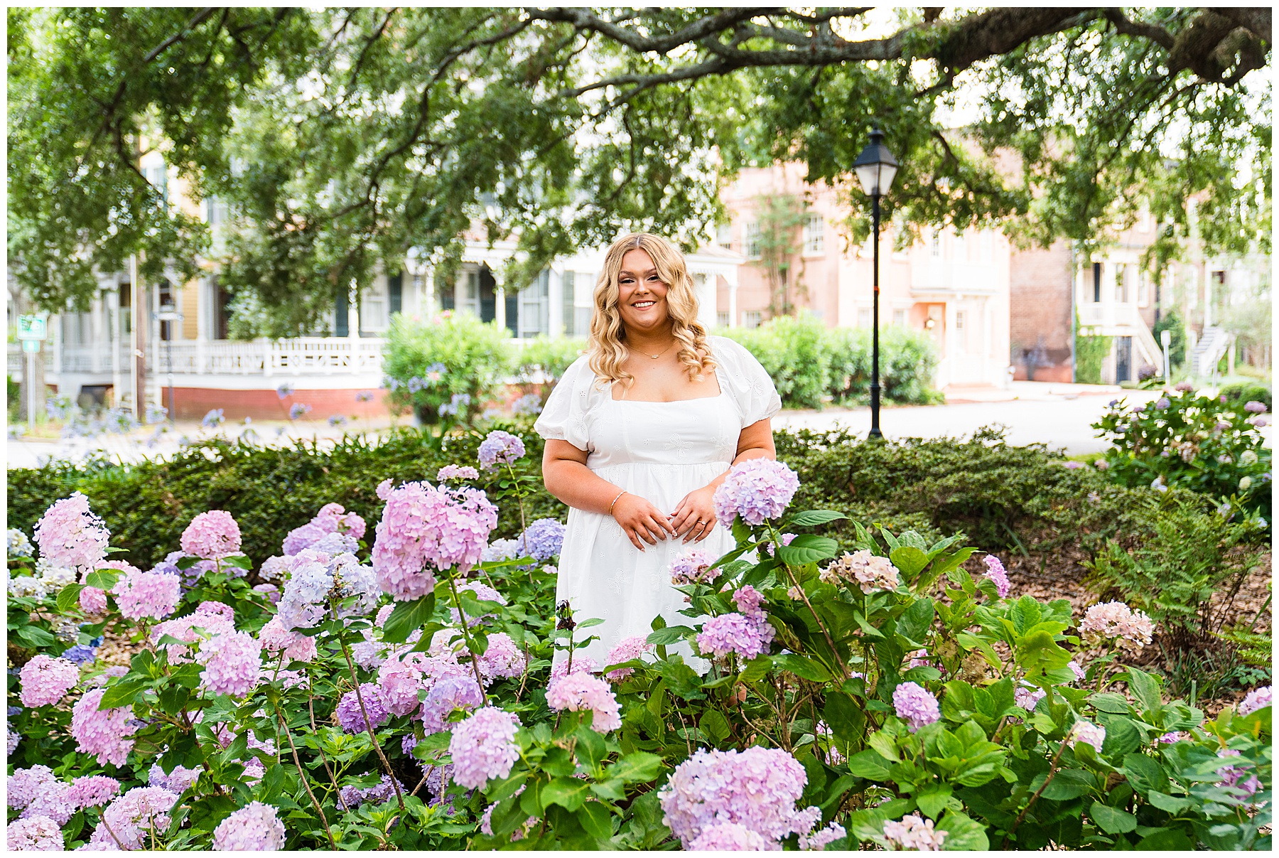 This senior portrait captures a soft and graceful moment in one of Savannah’s beautifully shaded squares: The senior is standing in the middle of a lush hydrangea garden filled with full, blooming clusters in pastel shades of pink, lavender, and pale blue. She’s wearing a white dress with subtle embroidery and puff sleeves, which gives off a fresh, timeless look that contrasts beautifully with the colorful flowers. Her hands rest gently in front of her, and she’s smiling warmly at the camera. Her long, loose curls cascade over her shoulders, perfectly styled for the natural setting. In the background, you can see classic Savannah architecture with porches, shutters, and soft pastel tones, along with a traditional streetlamp and oak branches draped in Spanish moss above. The composition is romantic and garden-inspired, with the senior beautifully centered and framed by blooms and greenery.
