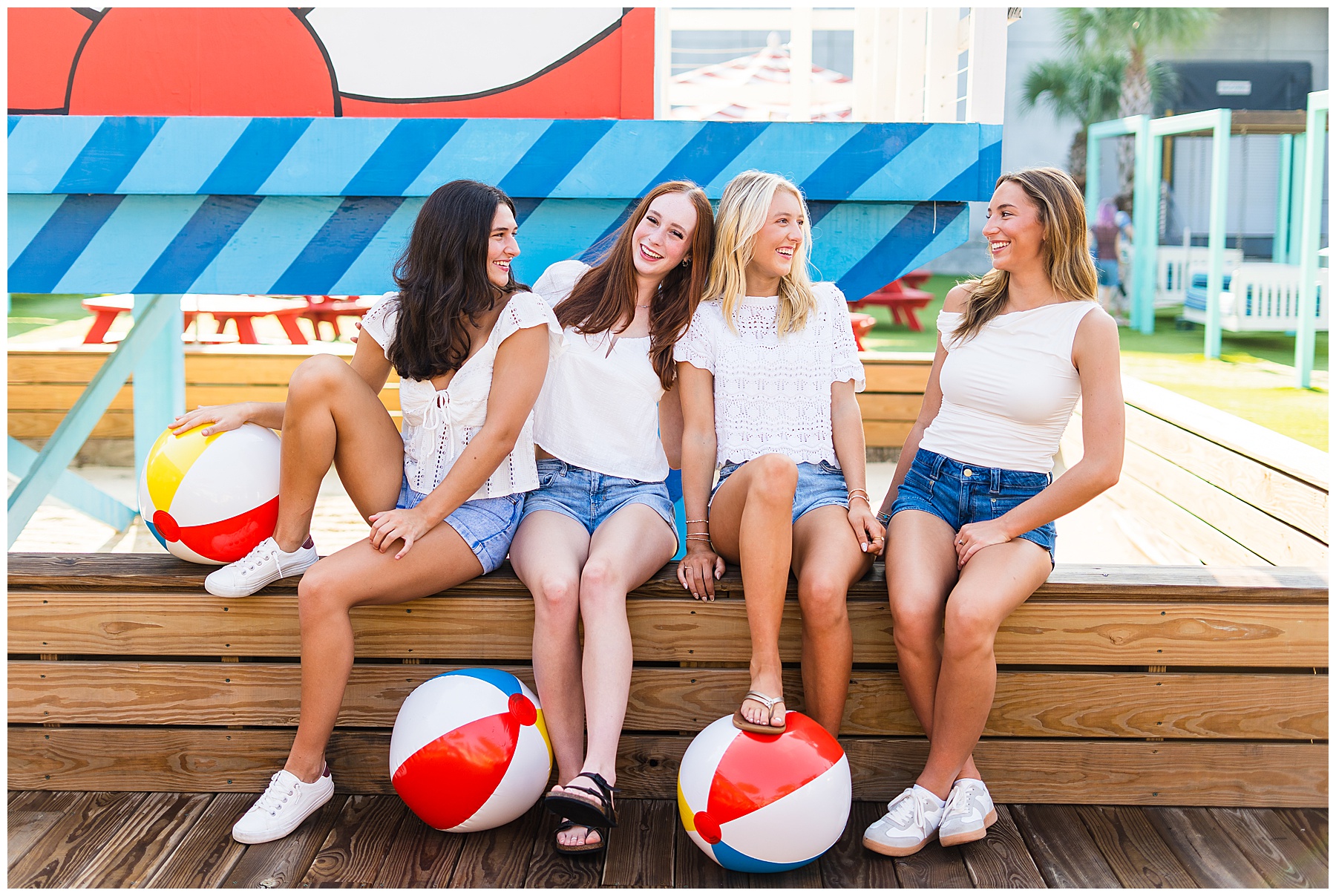 This photo captures a warm, candid group moment between the four young women. Scene & Composition The friends are seated side by side on a wooden ledge, leaning comfortably against each other while smiling and laughing. Each girl is dressed in coordinating outfits—white tops with denim shorts—maintaining the casual, summery theme carried throughout the series. Colorful beach balls rest at their feet and in their laps, adding a playful, lighthearted element to the scene. Setting Behind them, a structure painted in bright blue and red stripes adds a bold pop of color. The setting retains the lively, outdoor recreational feel of the earlier images, with picnic tables, palm trees, and sunshine visible in the background. Mood The overall vibe is joyful and carefree, emphasizing friendship and fun. The way the women are laughing and looking at each other highlights genuine connection, making this image feel natural and spirited rather than posed.
