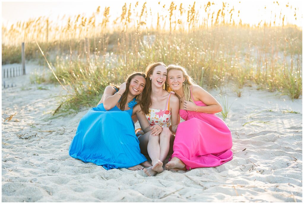 This image beautifully captures a relaxed, joyful beach moment among three young women.
Scene & Composition
The three friends are sitting closely together on sandy dunes, leaning into one another with warm, genuine smiles.
Their bright, colorful dresses — one in vibrant blue, one in floral, and one in hot pink — pop against the soft, golden tones of the beach backdrop.
They are barefoot, which adds to the carefree, natural feel of the photograph.
Background
The scene is set against tall sea oats swaying gently in the breeze, with the warm glow of the setting (or rising) sun casting a golden light across the frame.
The sunlit grasses add a dreamy, ethereal quality that enhances the warmth and intimacy of the image.
Mood
The overall feeling is serene, joyful, and deeply connected.
The way the women lean on one another conveys friendship, closeness, and an easy comfort.
The combination of playful colors, natural setting, and soft lighting makes this photo feel like a celebration of friendship and summer.