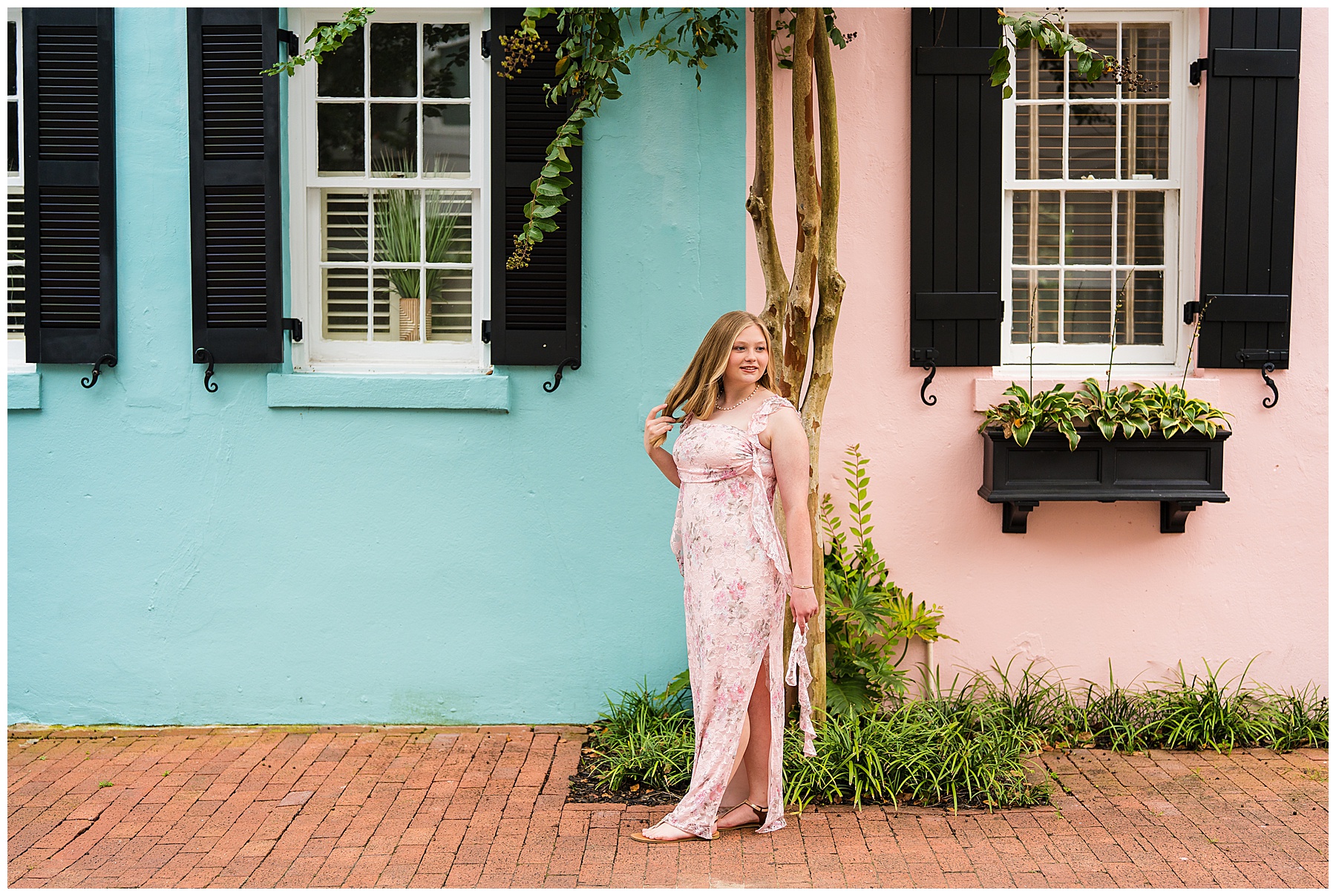 The image shows a young woman standing outdoors beside a pastel-colored building. The wall behind her is divided into two sections: the left side is painted turquoise blue, and the right side is light pink. Both sections have white-framed windows with black shutters. The pink side features a black flower box filled with green plants. A slender tree with light brown bark stands at the junction of the two wall colors, with some green leaves and small clusters of buds hanging down. The woman is wearing a light pink floral dress with a slit on one side and matching sandals. She has light hair and is smiling slightly, posing with one hand gently touching her hair. The ground is made of red brick, adding warmth to the scene. The overall mood is cheerful and summery, with a pleasing contrast between the pastel walls, black window details, and the woman’s floral outfit.