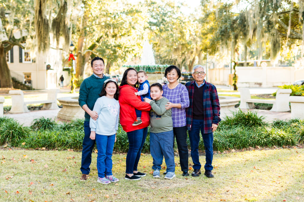 The image shows a multigenerational family posing together outdoors in a park-like setting. They are standing on grass in front of a fountain, with tall trees and hanging Spanish moss creating a picturesque background.
The group consists of seven people:
Two adults standing on the left, one with his arm around a young girl.
A woman in a bright red jacket holding a small child.
A boy standing next to an older woman, who has her arm around him.
An older man stands on the far right.
Everyone is smiling and appears relaxed and happy. The weather looks pleasant with soft sunlight filtering through the trees. The overall feeling of the image is warm, cheerful, and full of family togetherness.