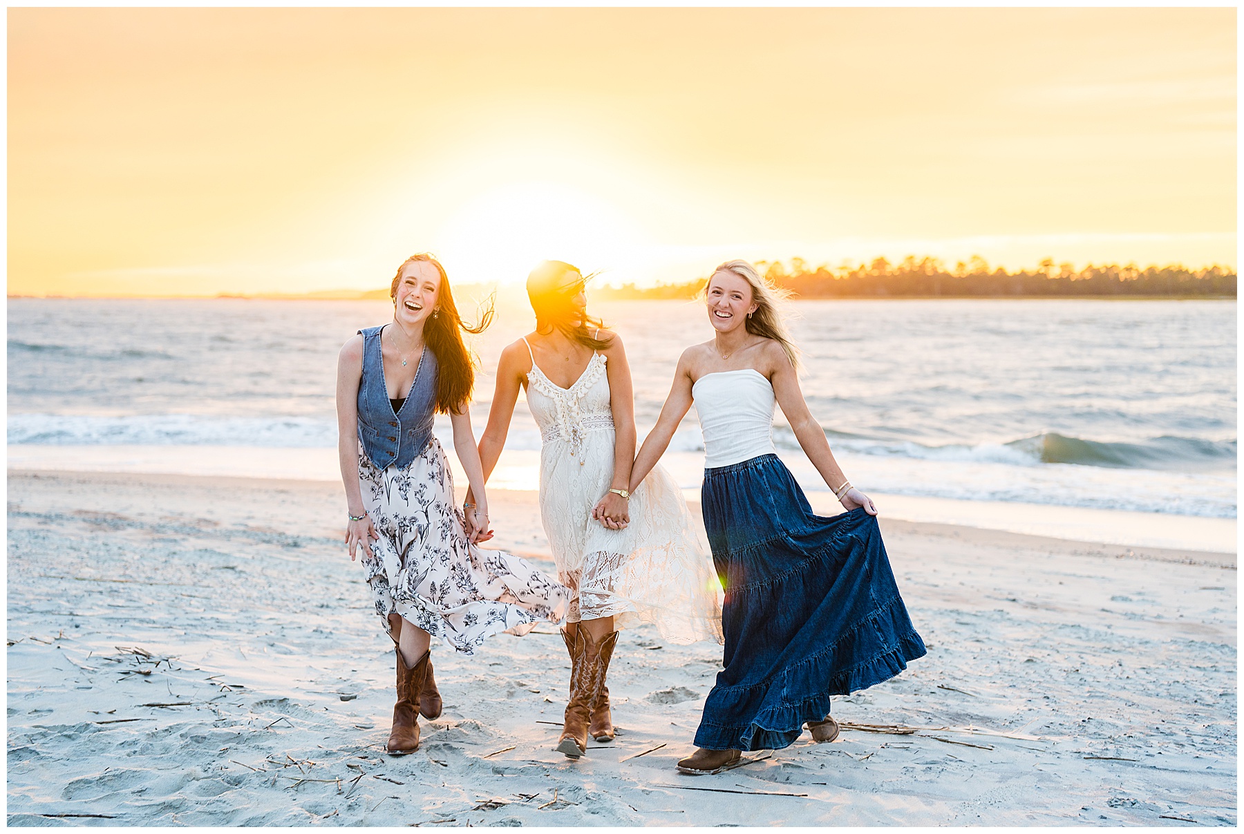 This image captures three young women walking hand in hand along a sandy beach at sunset, exuding happiness and friendship. They are all smiling and laughing as the golden sunlight shines behind them, creating a warm glow that silhouettes their figures and illuminates the waves in the background. Each woman is dressed in a flowy, bohemian-style outfit with a coastal flair — light dresses and long skirts paired with cowboy boots, perfectly suited for a casual yet stylish beach photoshoot. The woman on the left wears a floral dress with a denim vest, the one in the center is in a lacy cream dress, and the woman on the right has on a white strapless top with a long blue skirt. The setting sun, the soft motion of their skirts, and their carefree expressions all combine to create a joyful, radiant, and timeless scene that celebrates friendship and the beauty of a beach sunset.