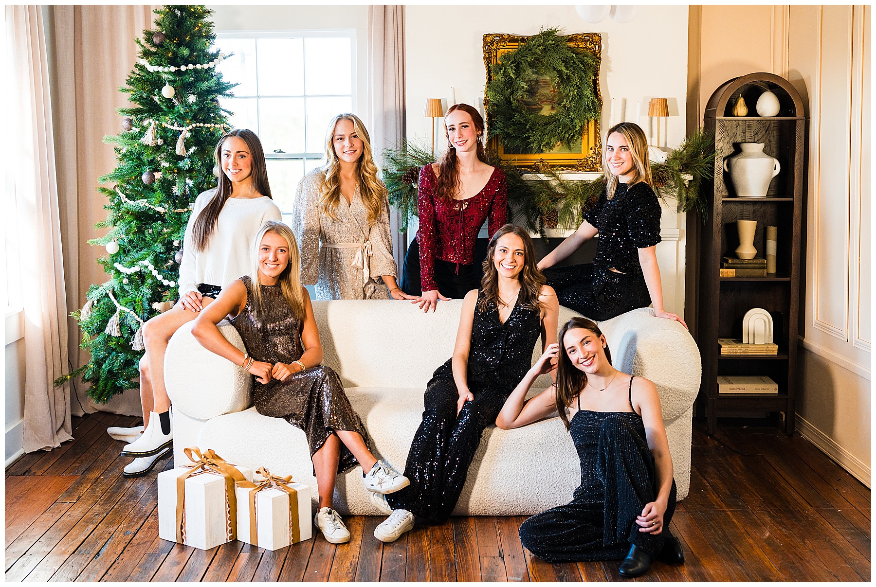 The image shows a festive group portrait taken indoors during the holiday season. Eight young women are posed together around a modern white sofa in a warmly decorated living room. They are dressed in semi-formal to dressy outfits—several wearing sequined dresses or jumpsuits in black, silver, and deep red—creating an elegant, celebratory feel. A decorated Christmas tree stands on the left side of the room with white and neutral-toned ornaments. Behind the group, a mantel is adorned with greenery, and a framed painting above it is accented with a large wreath. Soft, neutral décor and warm lighting give the space a cozy, stylish atmosphere. Some of the women are seated on the sofa, others lean or sit on the armrests, and one sits on the floor in front, all smiling and relaxed. Wrapped gift boxes with ribbon sit on the wooden floor near the sofa, reinforcing the holiday theme. Overall, the image feels like a polished holiday photo shoot capturing friendship, celebration, and seasonal warmth.