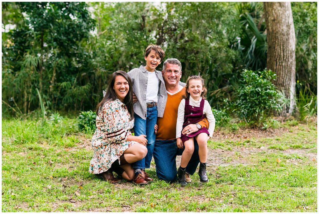 This image shows a family of four posed together outdoors in a lush, wooded setting, with a warm, candid, joy-filled feel. The family is gathered closely in the center of the image, kneeling and leaning into one another while laughing and smiling naturally at the camera. The background is filled with dense greenery, tall trees, and tropical foliage, creating a rich, natural backdrop that feels peaceful and secluded. Parents: The mother kneels on the left, smiling broadly. She has long dark hair and is wearing a cream floral dress with earthy tones paired with brown boots. The father kneels in the center-right wearing an orange/rust-colored sweater layered over a white shirt with dark jeans, smiling warmly. Children: Their son stands between the parents, slightly elevated and leaning in with an energetic expression. He wears blue jeans, a white shirt, and a lightweight checkered overshirt, giving a casual, classic look. Their daughter sits on her father’s knee, laughing brightly. She wears a deep burgundy pinafore-style dress over a cream long-sleeve top, paired with dark ankle boots. The styling feels cozy and autumn-inspired, with warm rust, burgundy, denim, and floral tones that complement the natural greens of the setting. Unlike a formal posed portrait, the image feels very genuine and personality-driven, capturing connection and laughter rather than stiffness. Overall, the photo has a warm, relaxed family storytelling aesthetic — polished but natural, with a focus on joy, closeness, and authentic expressions.